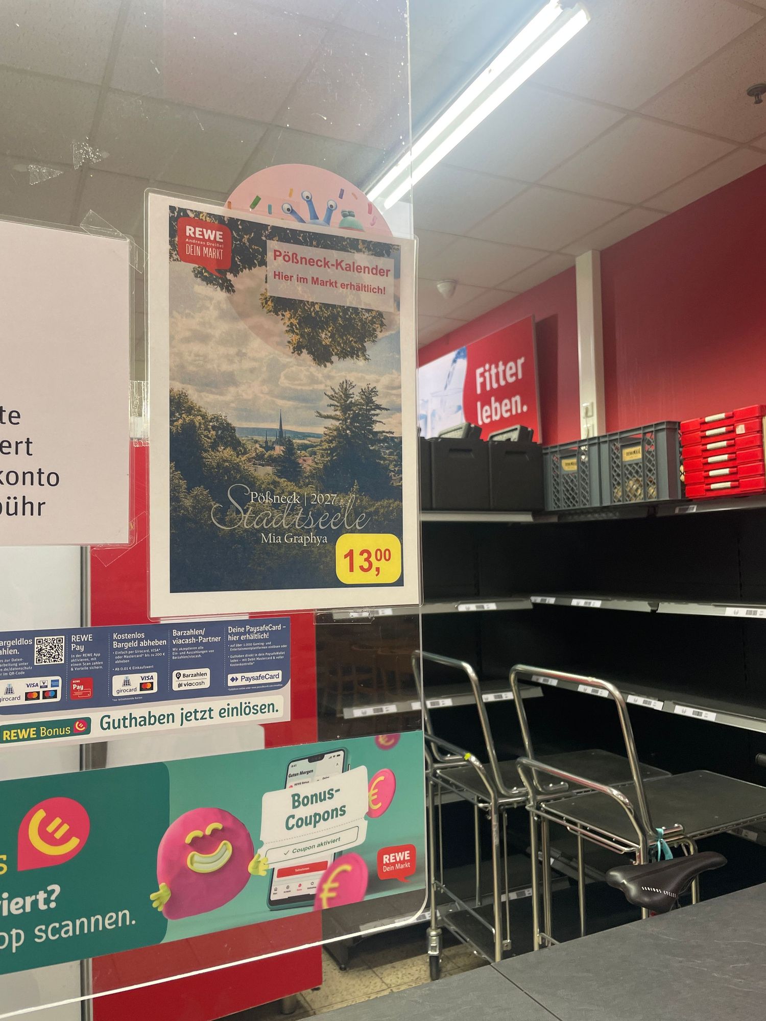 Interior of a REWE supermarket in Germany with empty shelves, promotional signs, and a calendar poster priced at 13.99 euros.