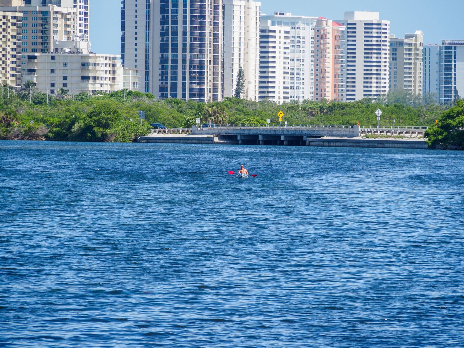 Kayaker paddling on blue urban waterway with modern high-rise buildings and bridge in background on sunny day.