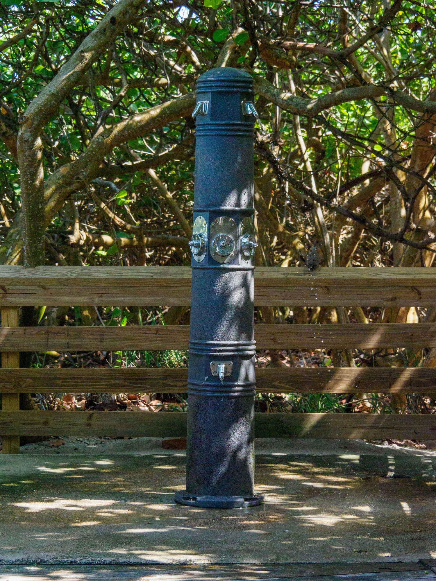 Dark cast iron decorative outdoor column post on wooden deck surrounded by lush green trees and wooden fence.