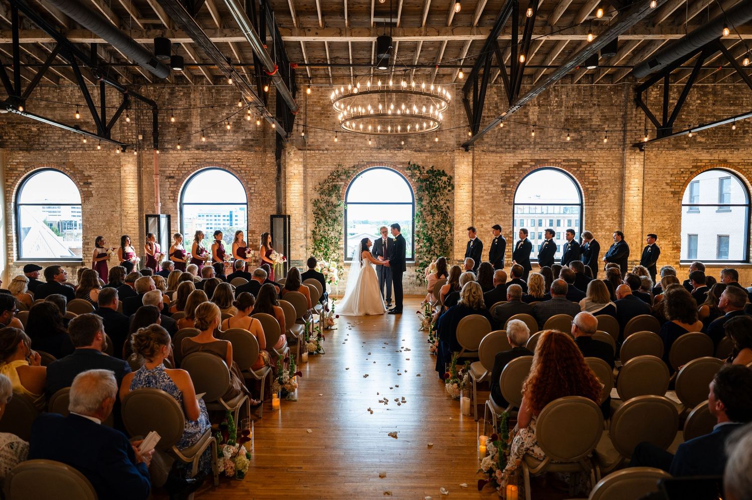 Elegant indoor wedding ceremony in a brick venue with chandeliers, floral arch, and guests seated in rows.