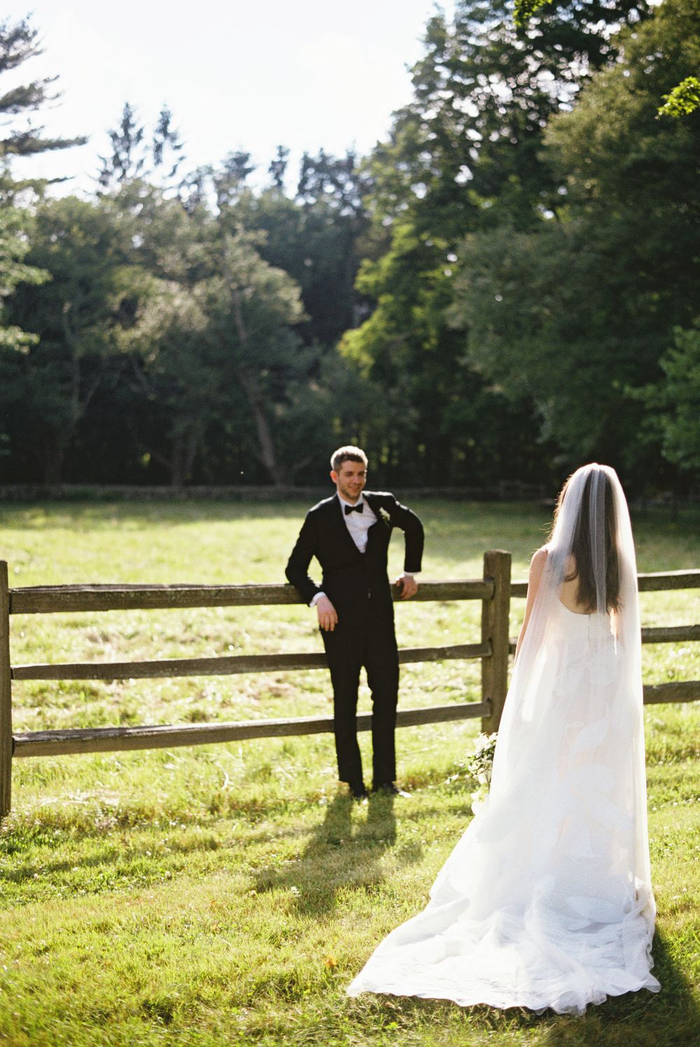 Bride in white gown and veil approaches groom in tuxedo by rustic wooden fence in lush green countryside.