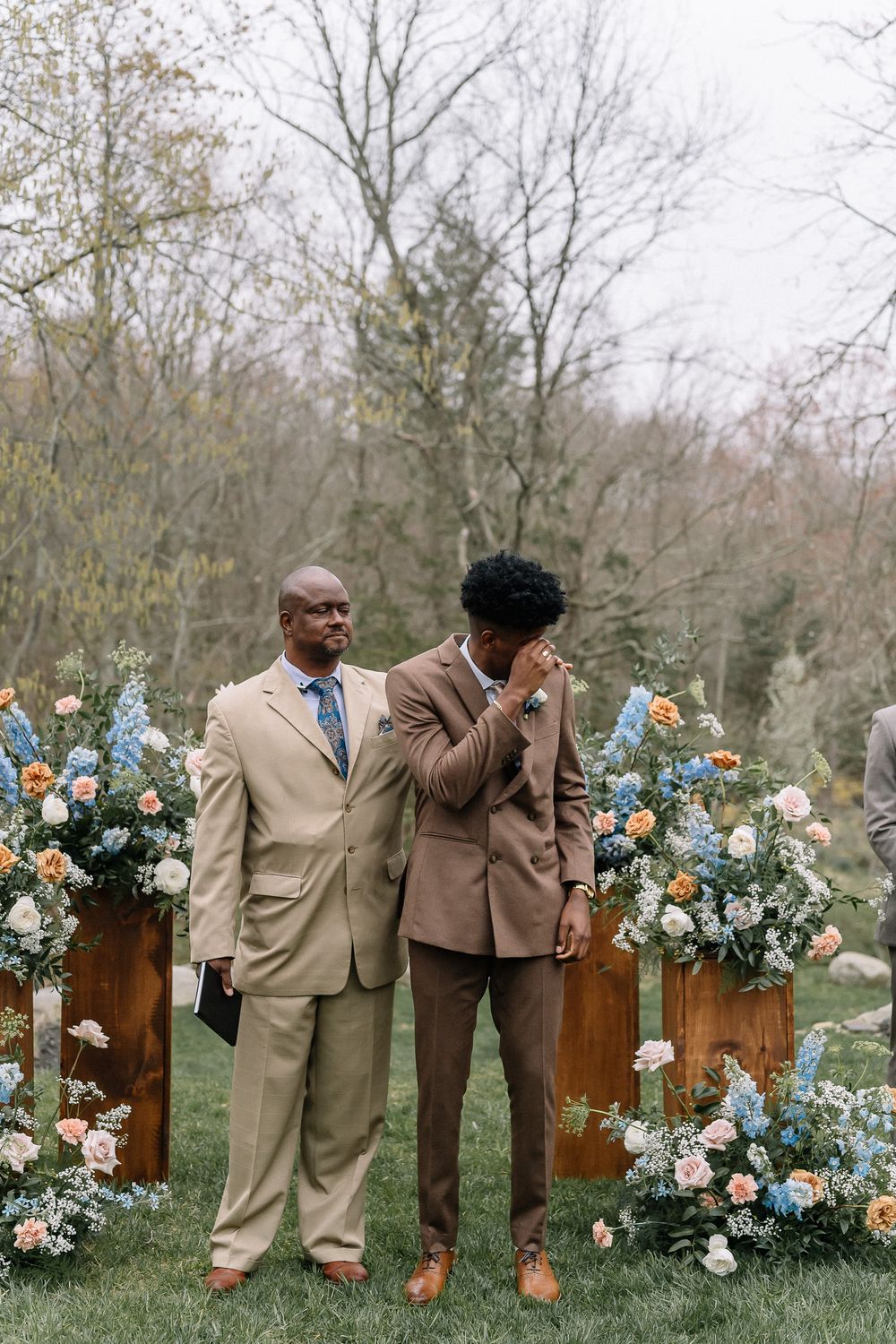 Groom wiping tears at outdoor wedding ceremony beside officiant, surrounded by lush floral arrangements in blue, peach, and white.