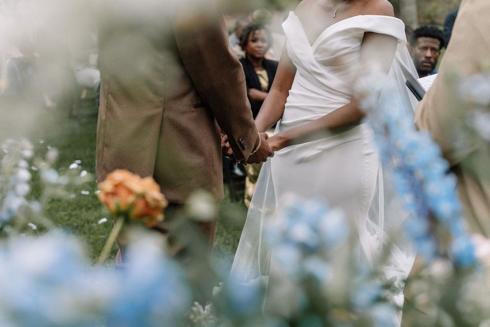 Bride in white off-shoulder gown holds hands with groom at outdoor wedding ceremony surrounded by blue flowers.