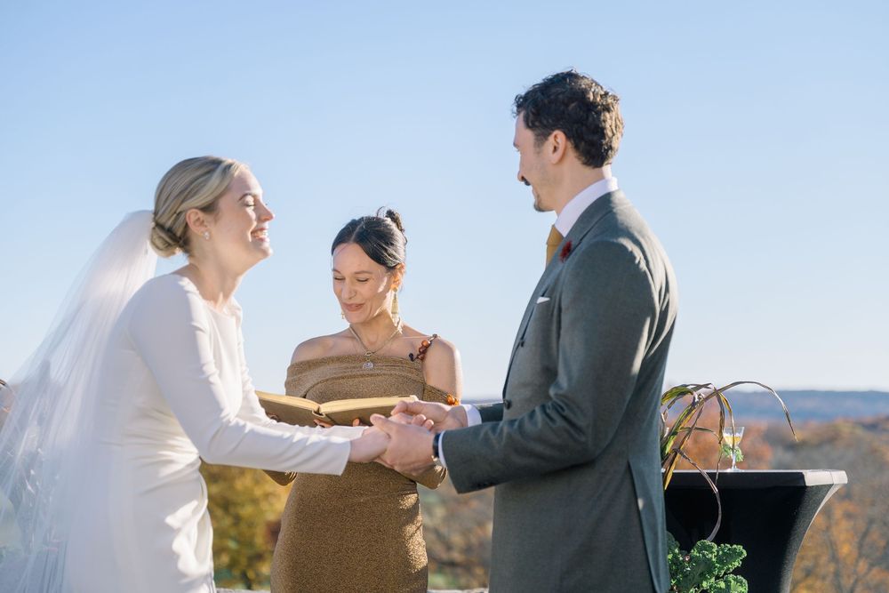 Bride and groom exchange vows outdoors with smiling officiant under clear blue sky at scenic hilltop wedding ceremony.
