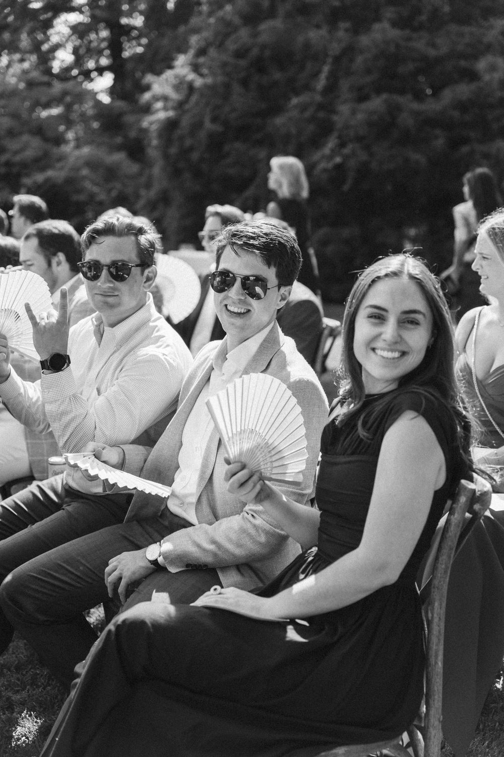 Black and white photo of smiling guests at an outdoor wedding, woman holding a hand fan, two men wearing sunglasses seated behind her.