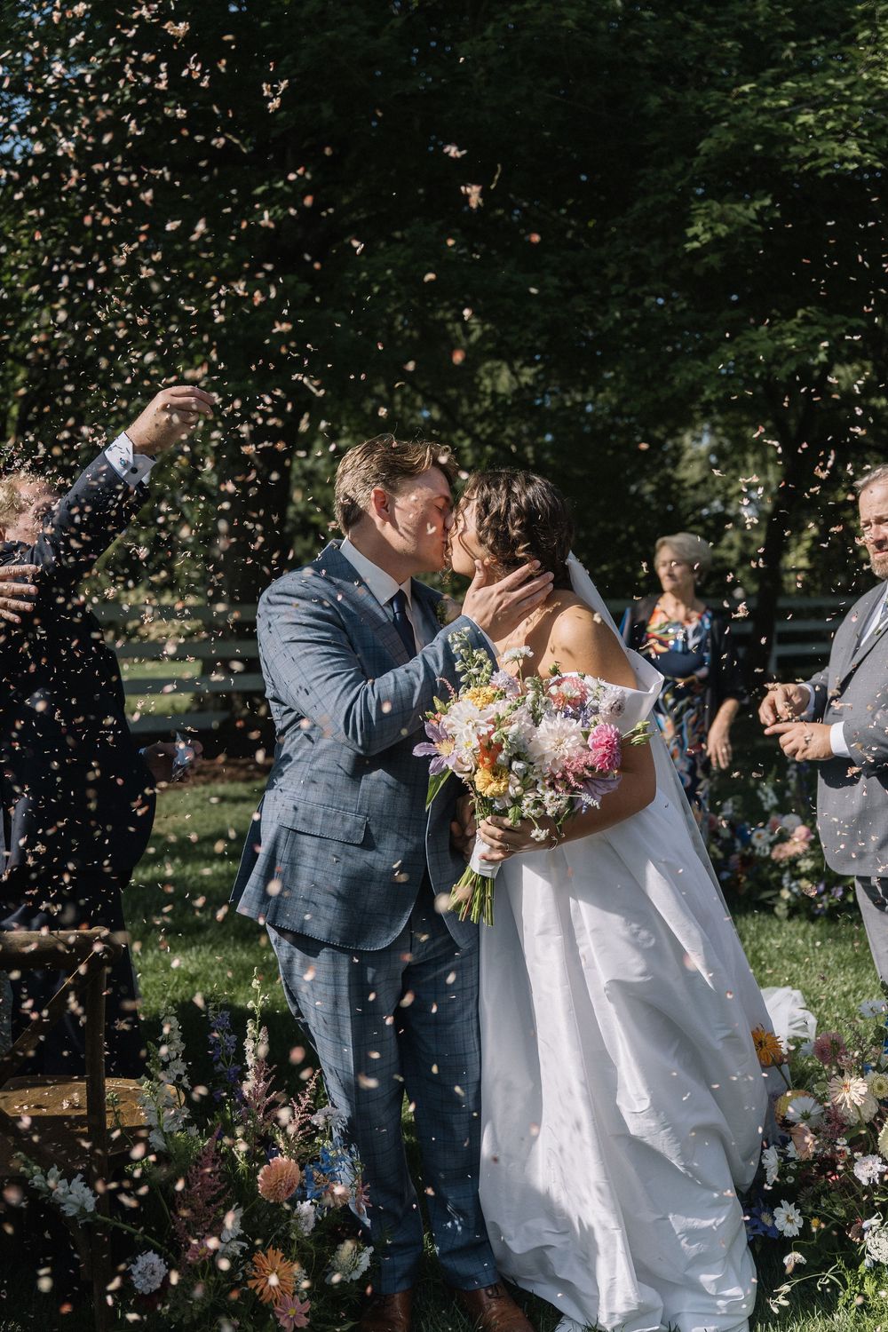 Bride and groom share a kiss during confetti shower at outdoor wedding ceremony, bride holding colorful wildflower bouquet.