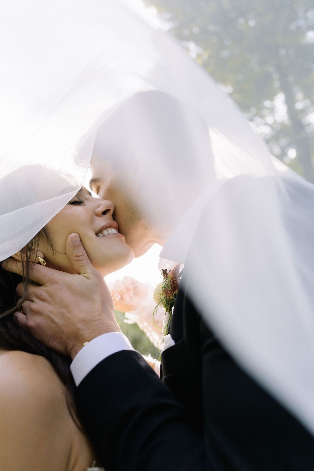 A bride and groom share a romantic kiss under a flowing white veil on their wedding day outdoors.