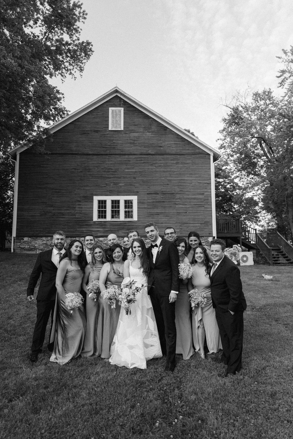 Black and white wedding party photo in front of rustic red barn with bride, groom, bridesmaids, and groomsmen.