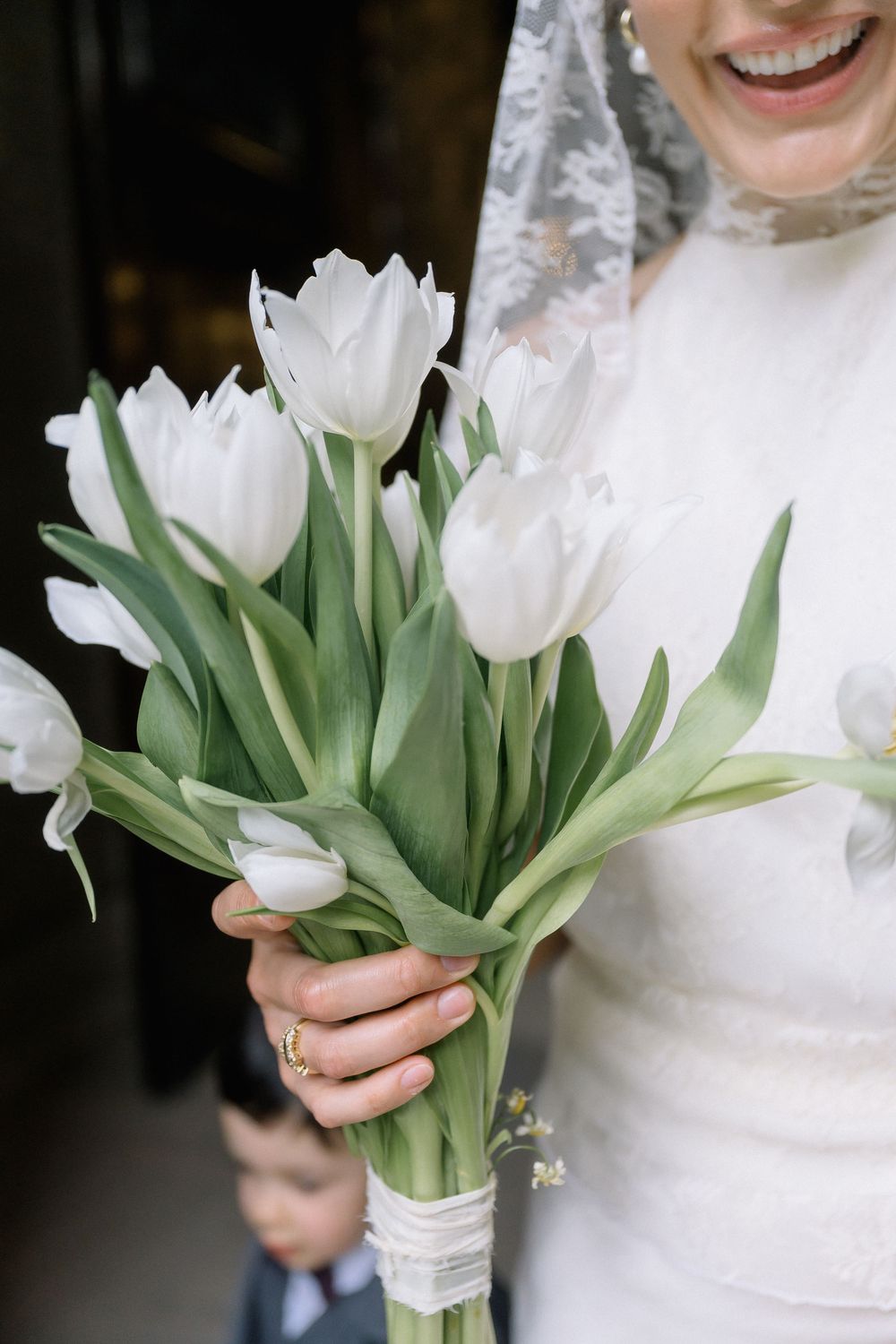 Bride holding a bouquet of white tulips wrapped in lace ribbon, wearing a white wedding dress and veil.