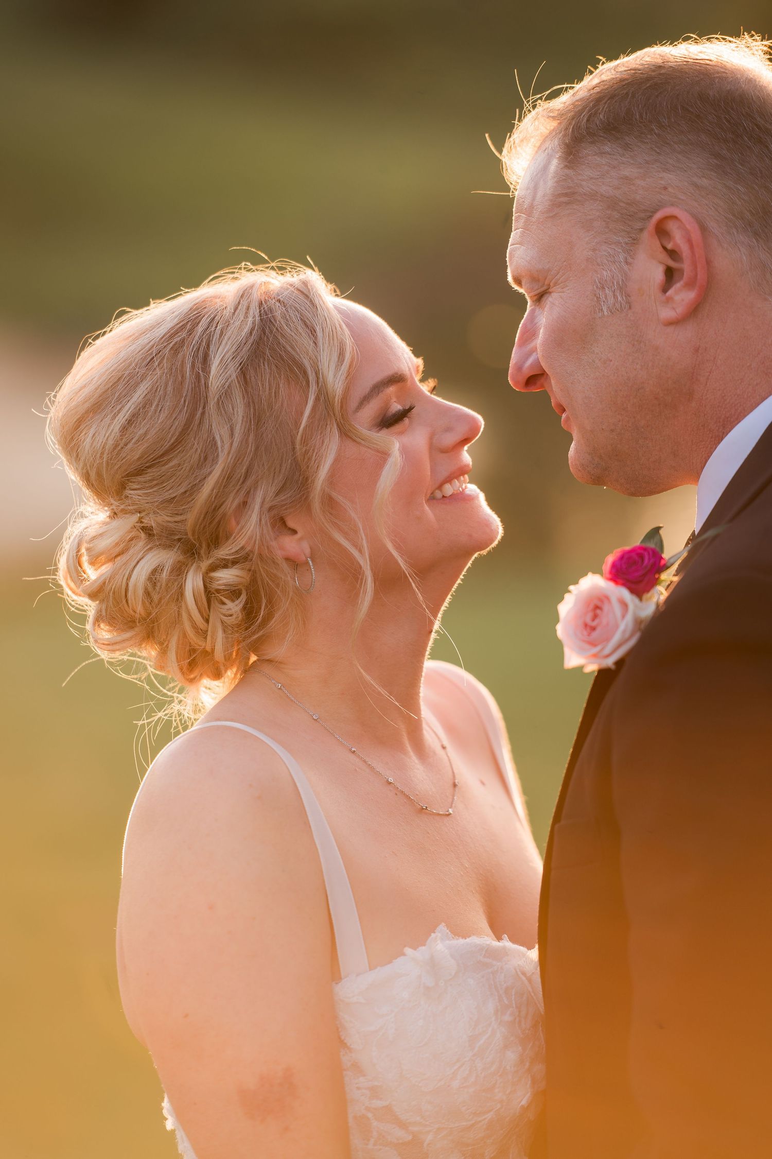A radiant bride with an updo smiles at her groom in golden hour light, both dressed in wedding attire with a floral boutonniere.