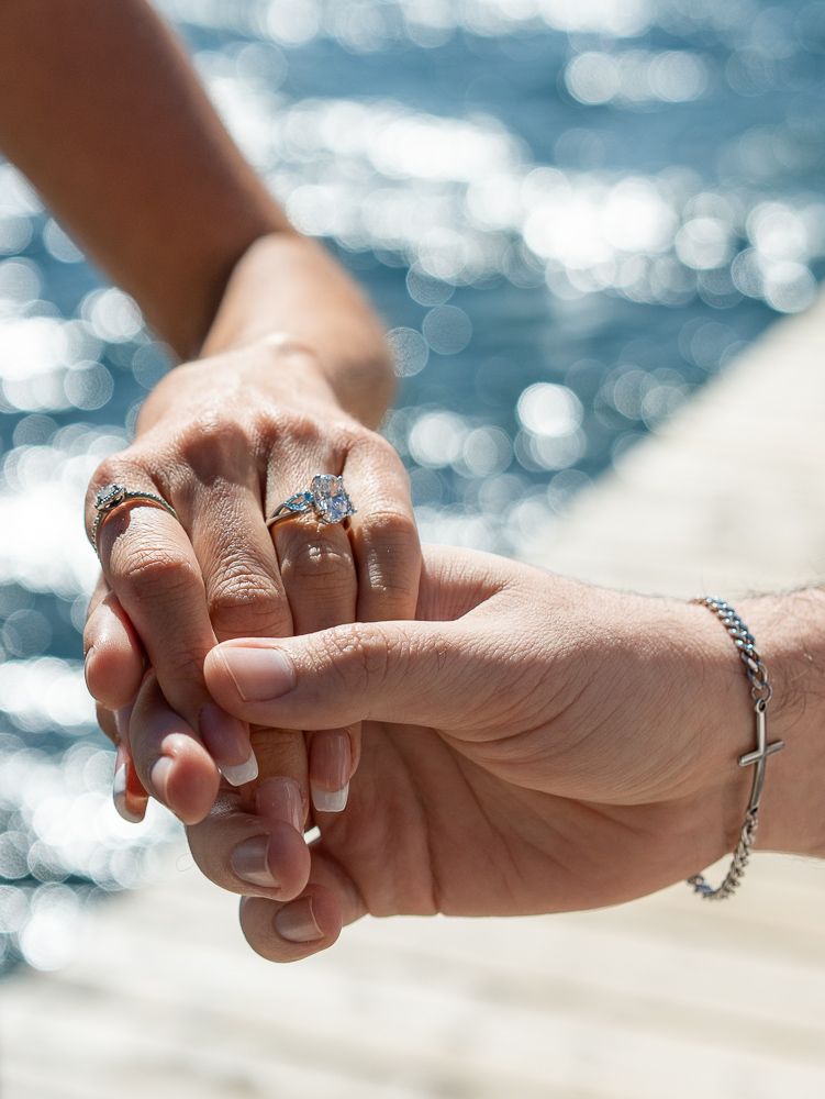 Two hands clasped together near sparkling water, adorned with diamond engagement rings and a silver bracelet.