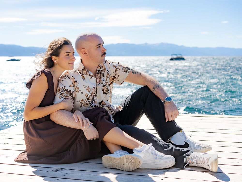 Couple sitting on a wooden dock by a sparkling blue lake, mountains in the background on a sunny day.