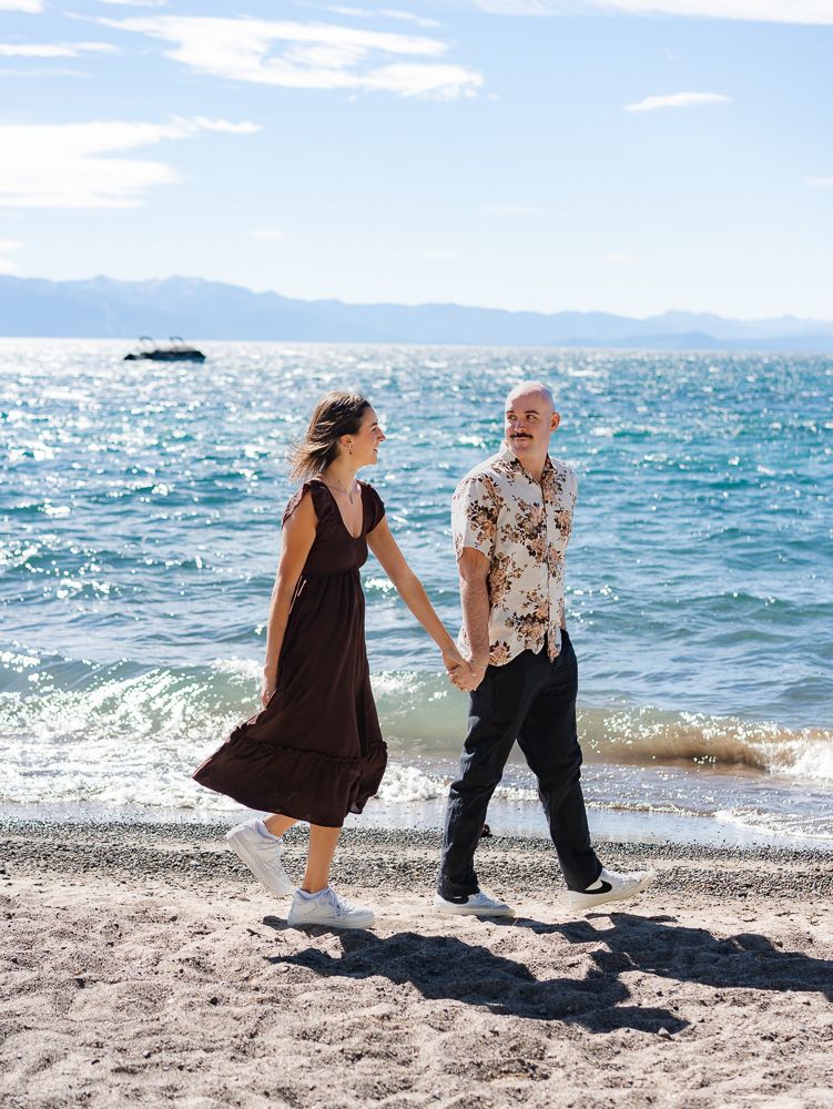 Couple holding hands and walking along a sunny sandy beach with turquoise lake water and mountains behind them.