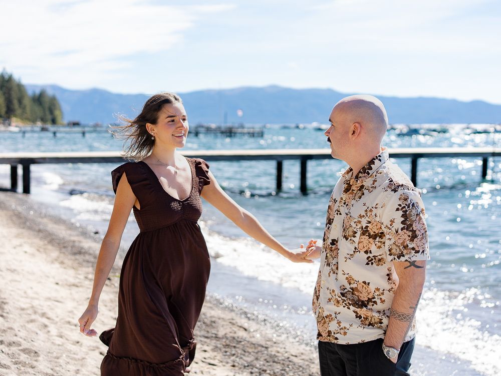 Pregnant woman in brown dress holding hands with bald man on a sunny lakeside beach with pier behind them.
