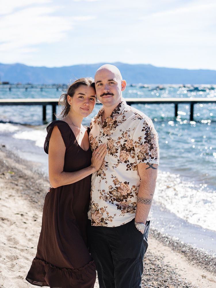 Couple embracing on a sunny beach with a pier and mountain lake backdrop, woman in brown dress, man in floral shirt.