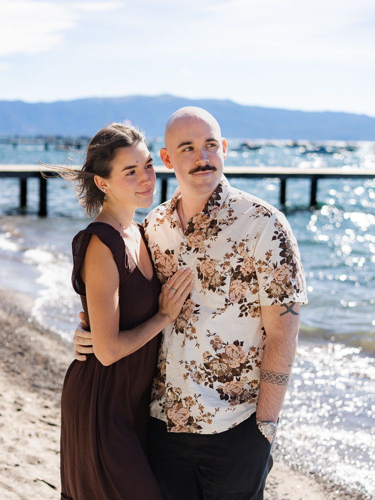 Couple posing on a lakeside beach, woman in brown maxi dress leaning on man in floral shirt, pier visible behind them.