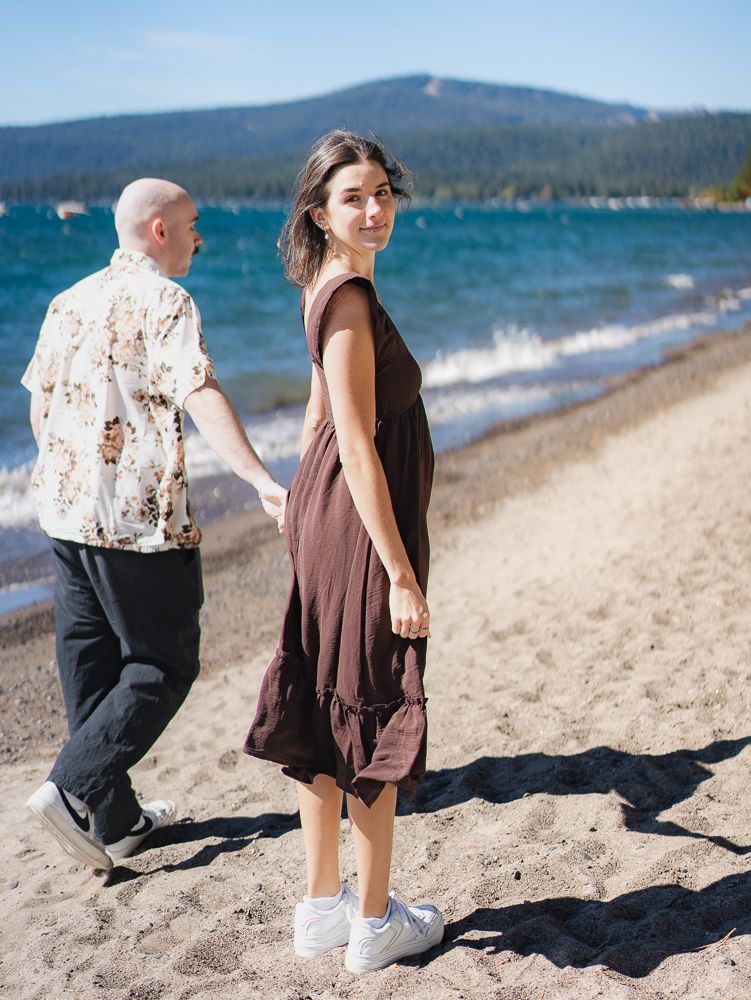 Woman in brown dress looks back smiling while holding hands with partner walking along a scenic mountain lake beach.