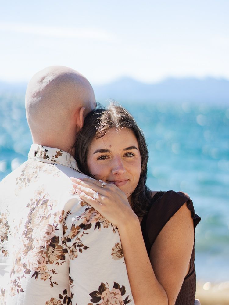 A couple embraces near a lake, woman in brown top smiles at camera while man in floral shirt holds her close.