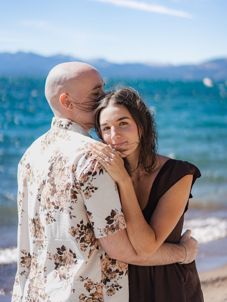 A couple embraces on a sunny beach near a blue lake with mountains in the background.
