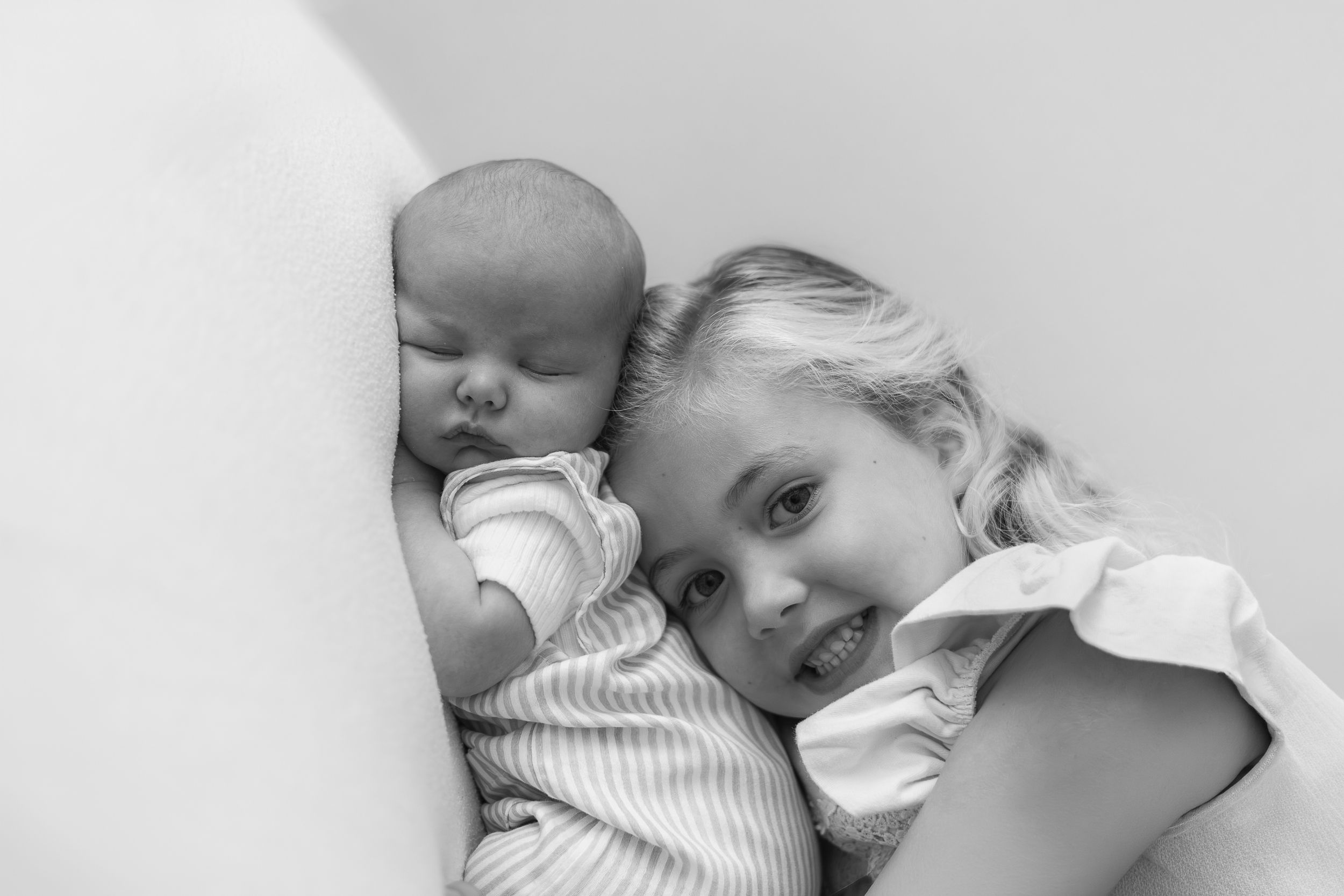 Black and white photo of a smiling girl lying next to a sleeping newborn baby sibling.