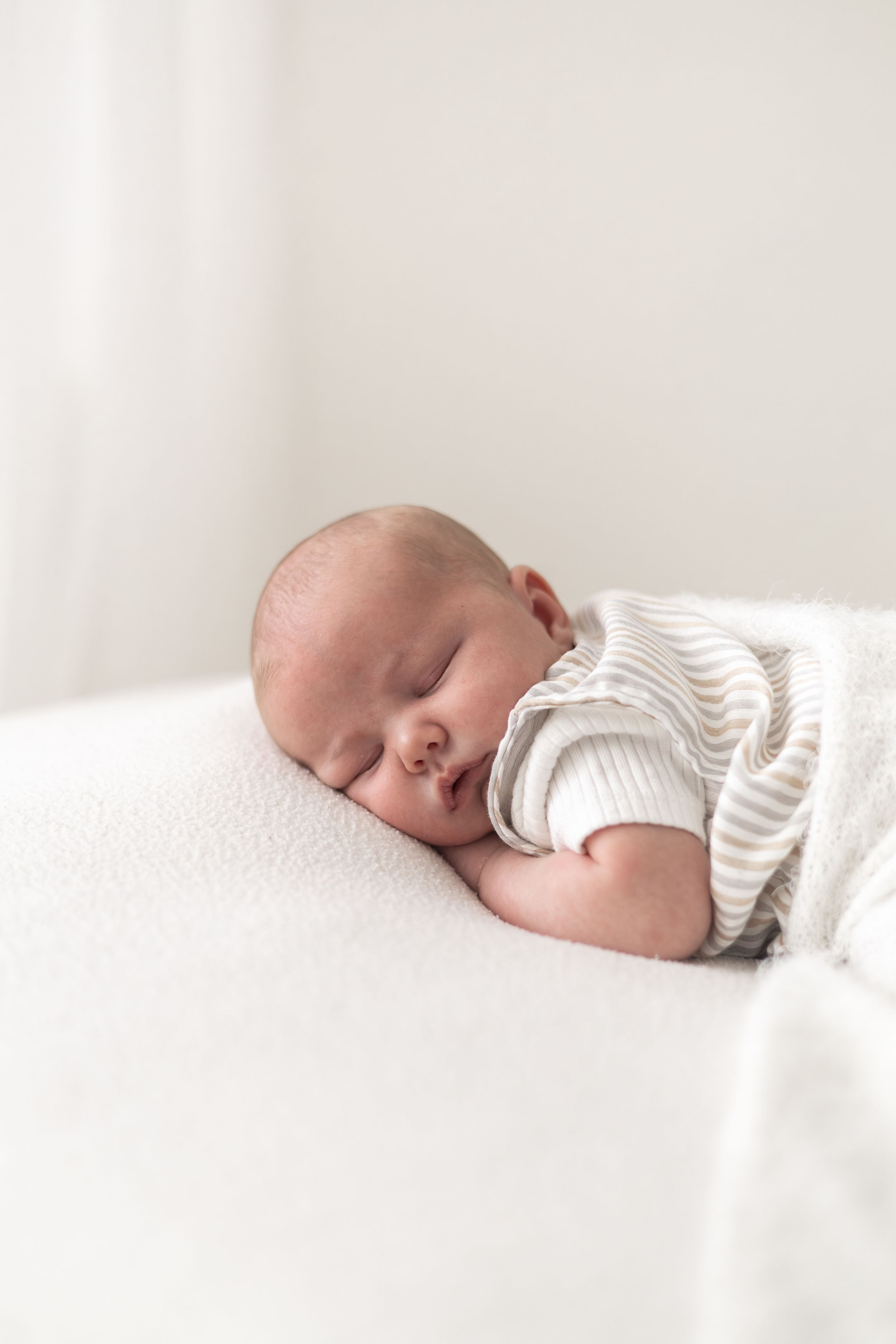 Peaceful newborn baby sleeping on a soft white blanket wearing a striped onesie in a bright, minimalist setting.