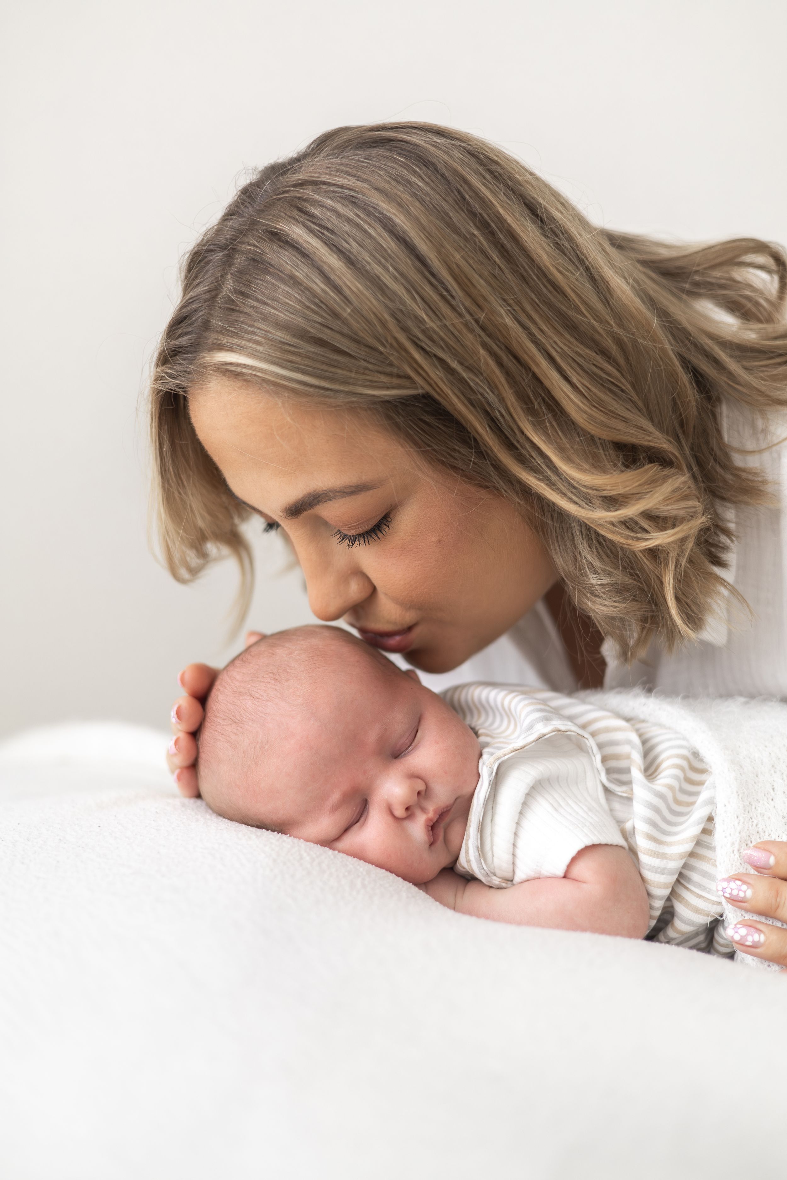 A young mother gently kisses her sleeping newborn baby dressed in a striped onesie on a soft white blanket.