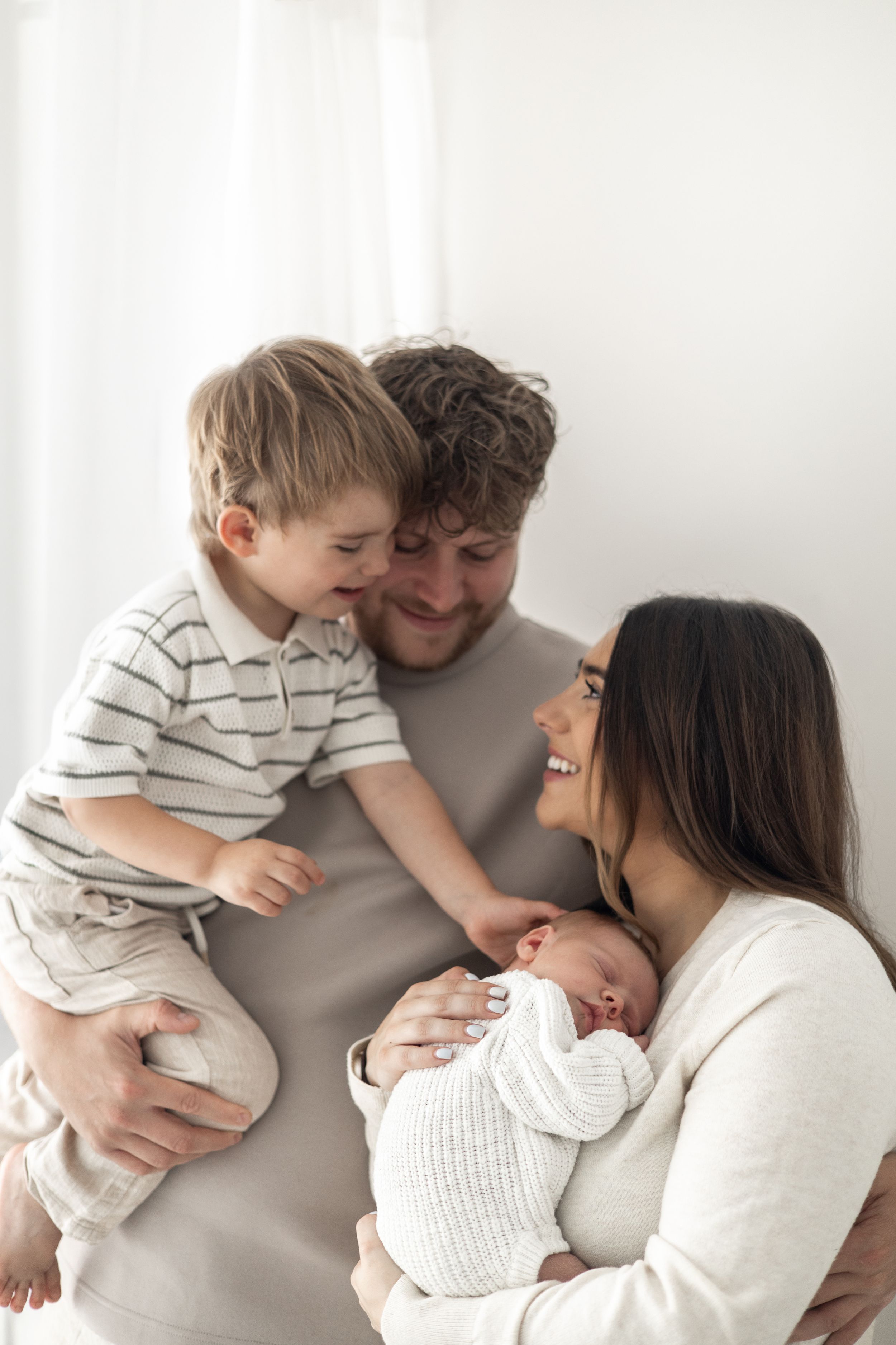 Happy family with father holding toddler son and mother cradling newborn baby, smiling together in bright white room.