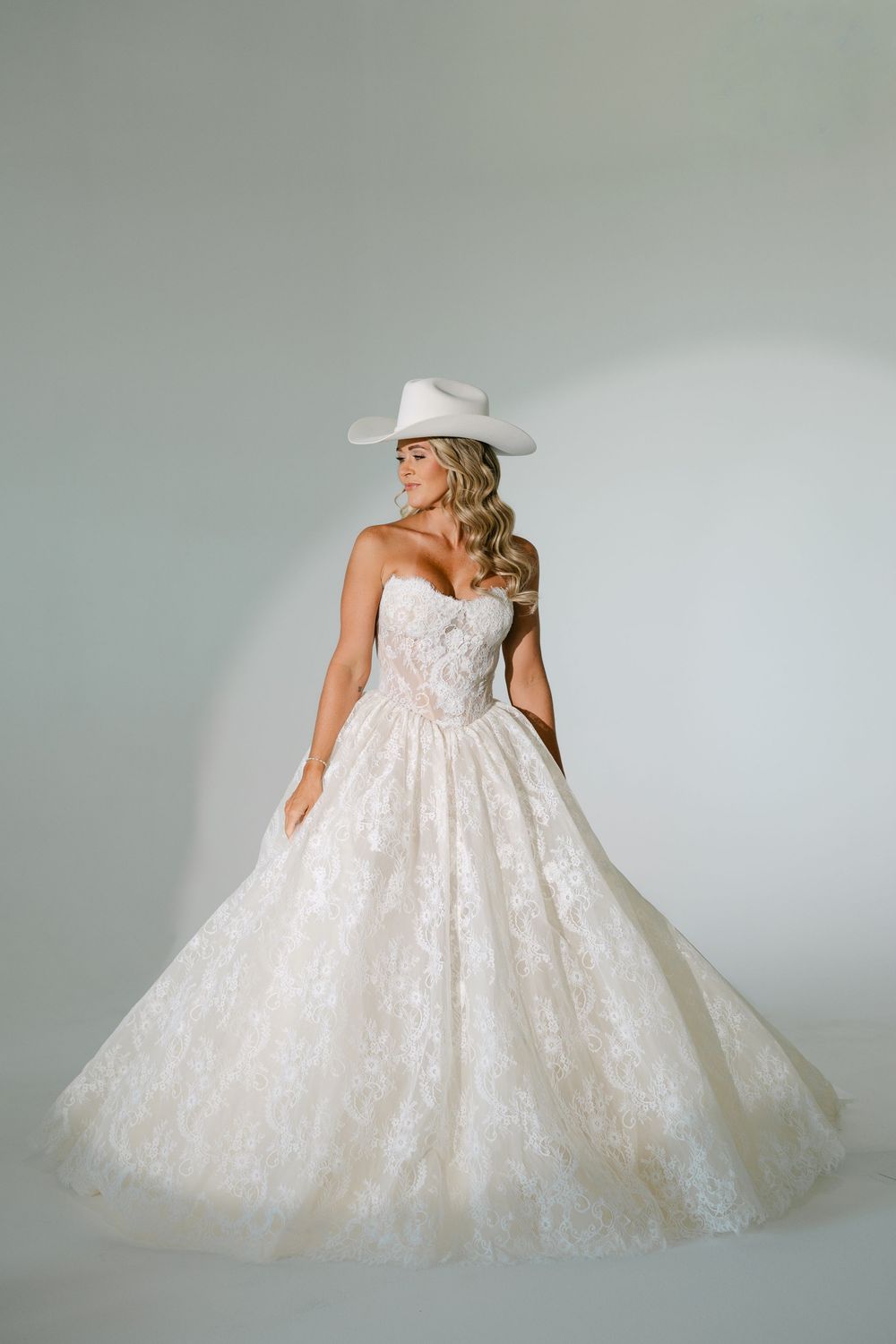 Bride in strapless lace ball gown wedding dress with white cowboy hat, posing elegantly in studio.