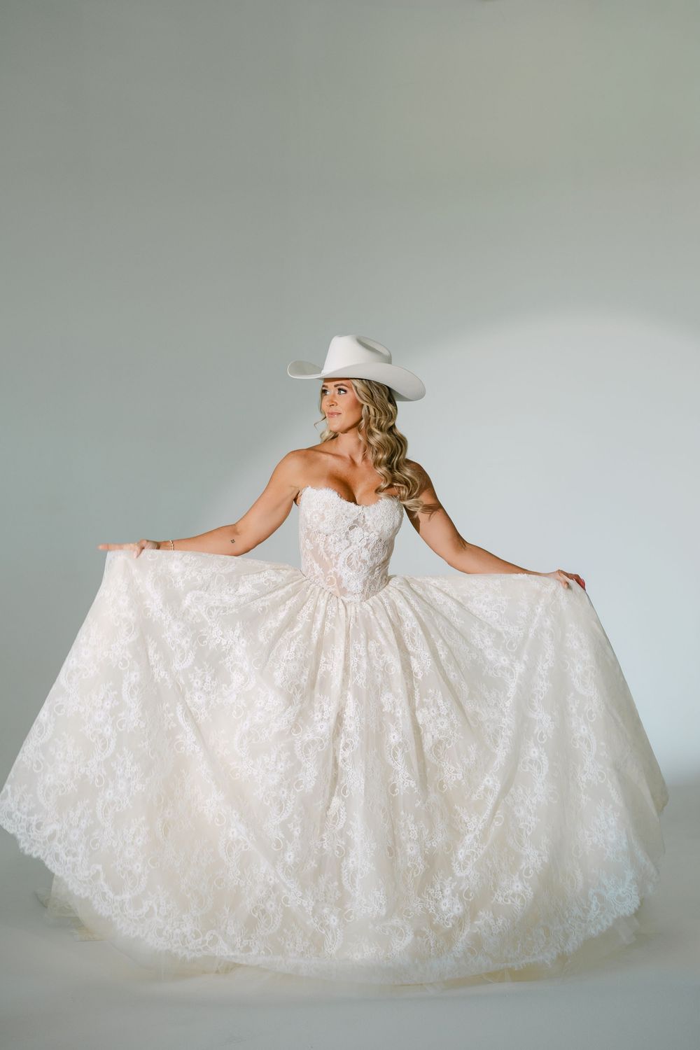 Bride in strapless lace ballgown wedding dress and white cowboy hat posing in studio.