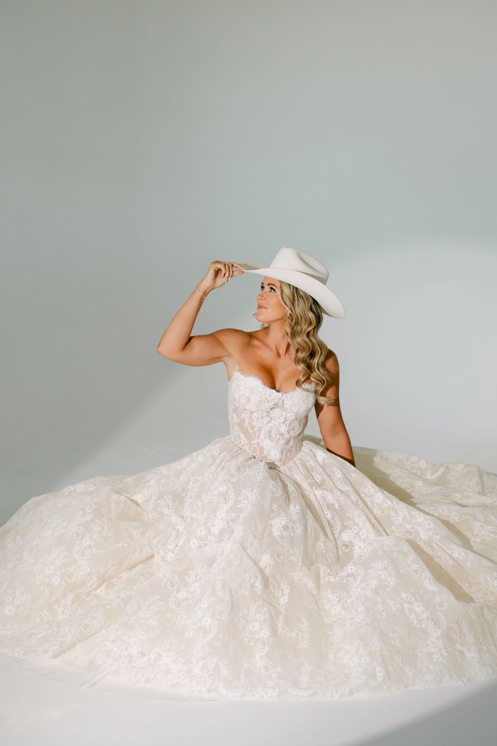 Bride in strapless lace ball gown and white cowboy hat sits elegantly against a soft gray background.