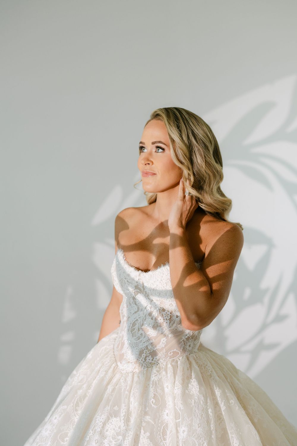 Bride in a strapless lace ballgown touches her neck, posing against a white wall with soft leaf shadows.
