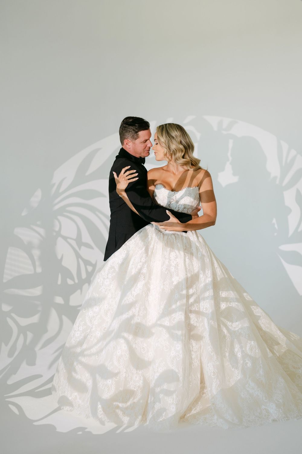 Bride and groom share a romantic embrace in a studio, she wearing an elegant lace ballgown wedding dress.