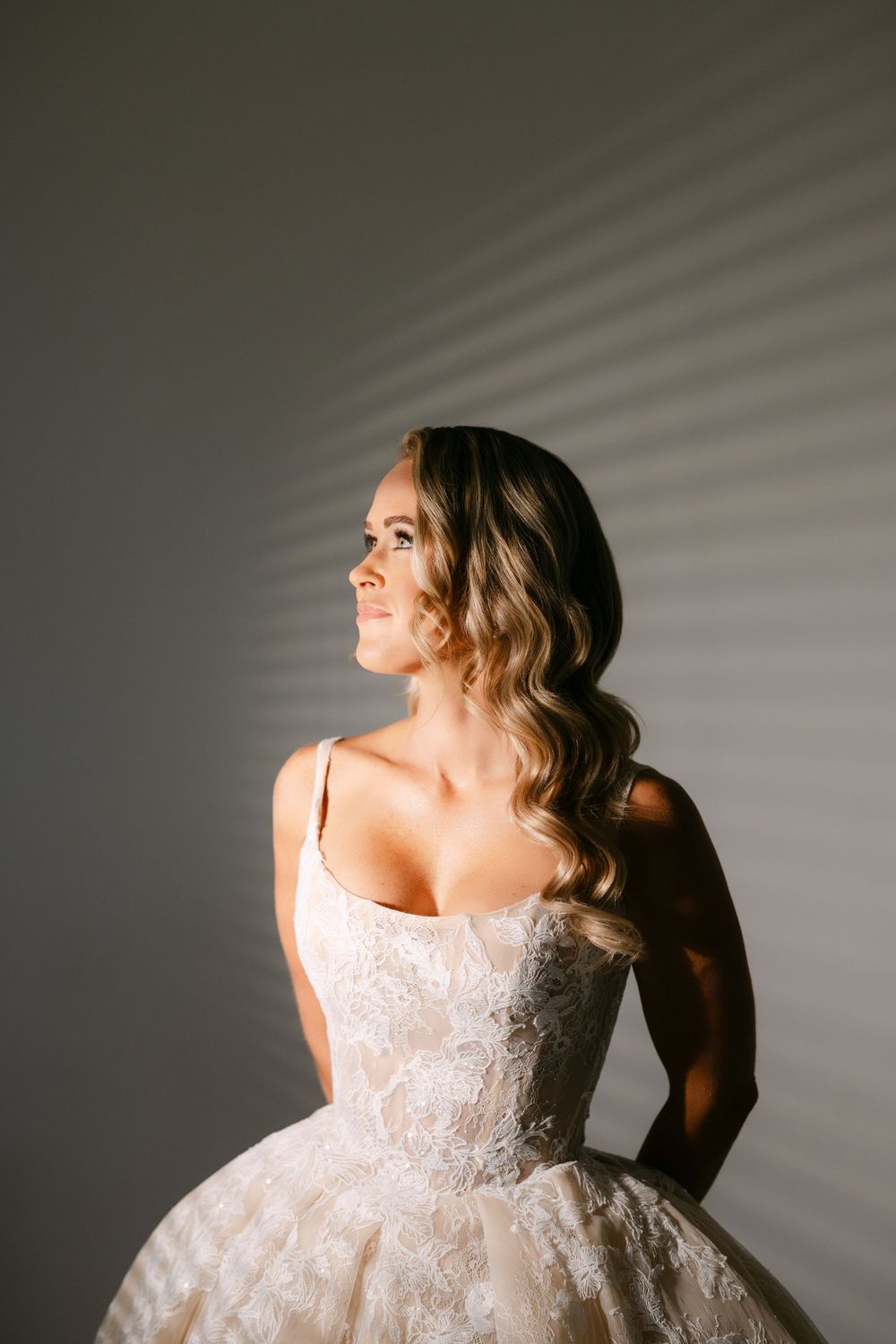 Bride in lace ballgown smiling in soft light with wavy hair.