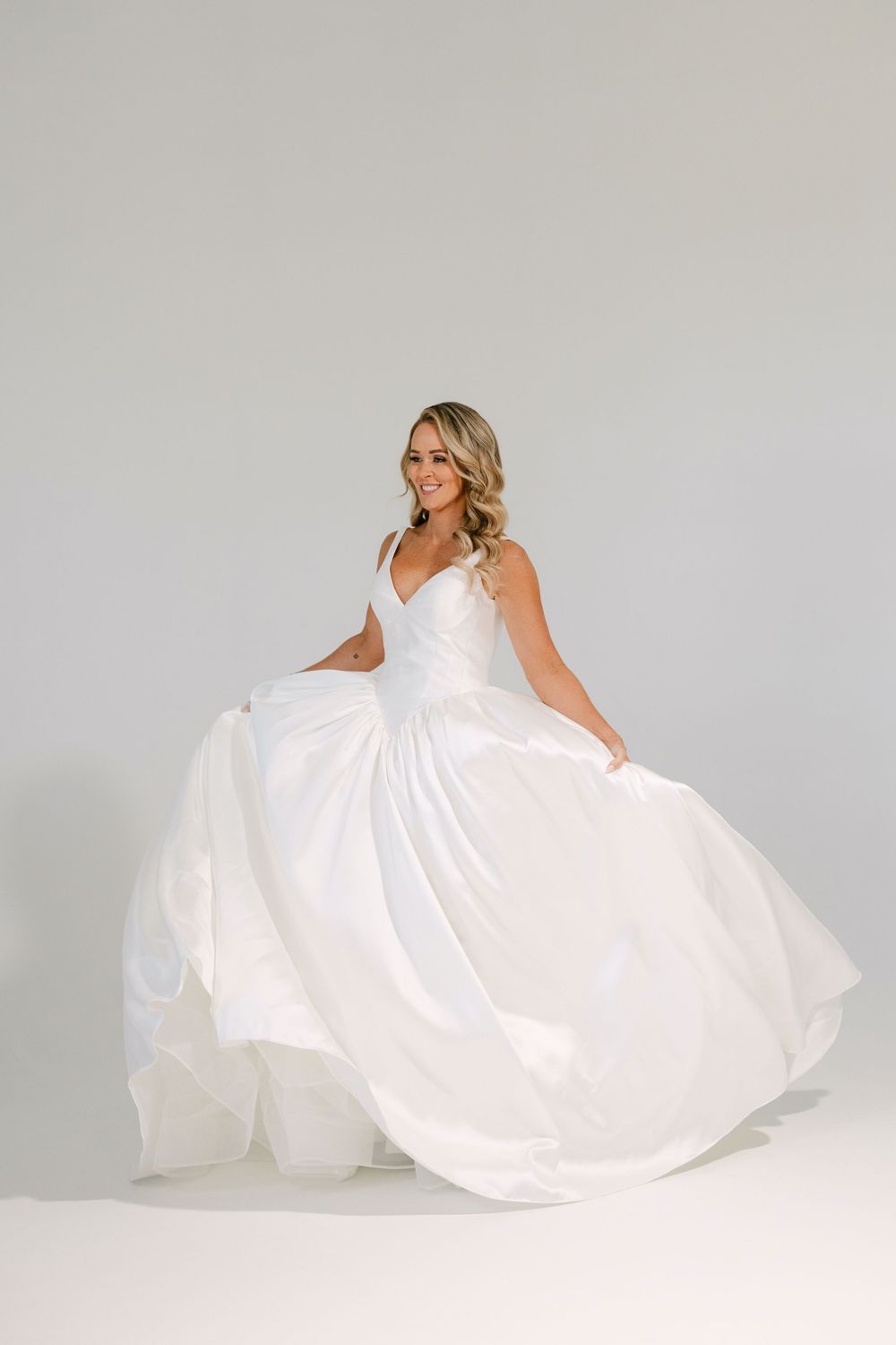 Smiling bride twirling in a flowing white ballgown wedding dress against a soft gray studio background.