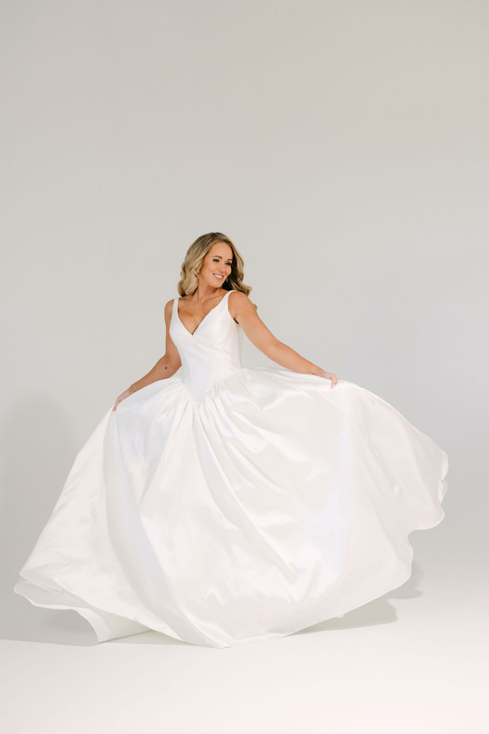 Bride twirling in a flowing white ball gown with V-neckline on a light gray background.
