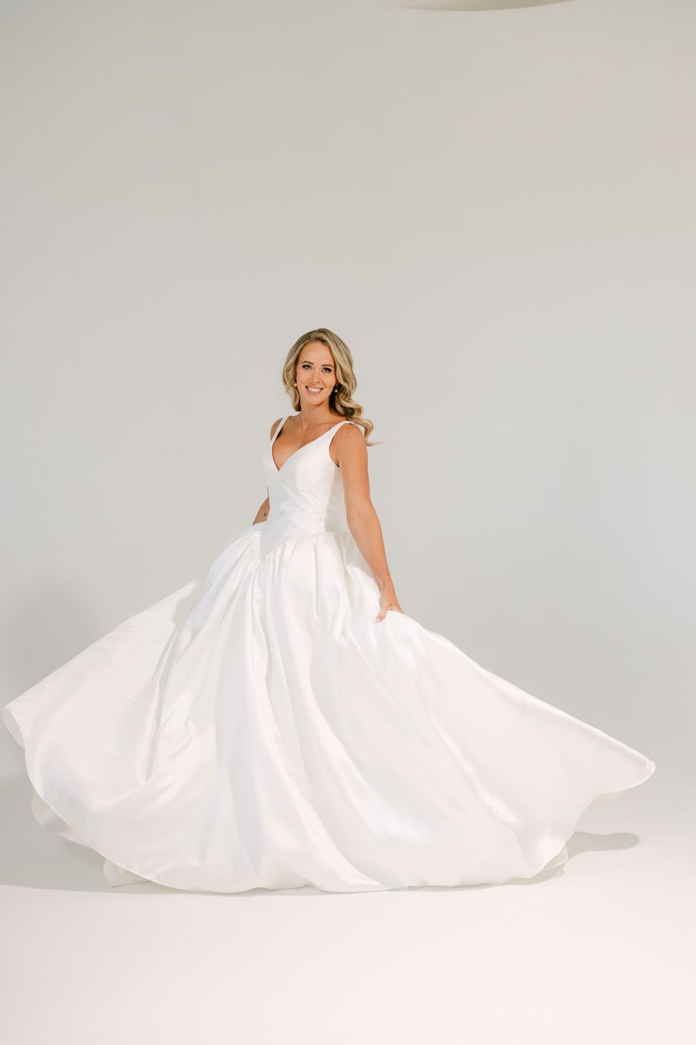 Bride twirling in a flowing white ballgown wedding dress with V-neckline and full skirt in a bright studio.