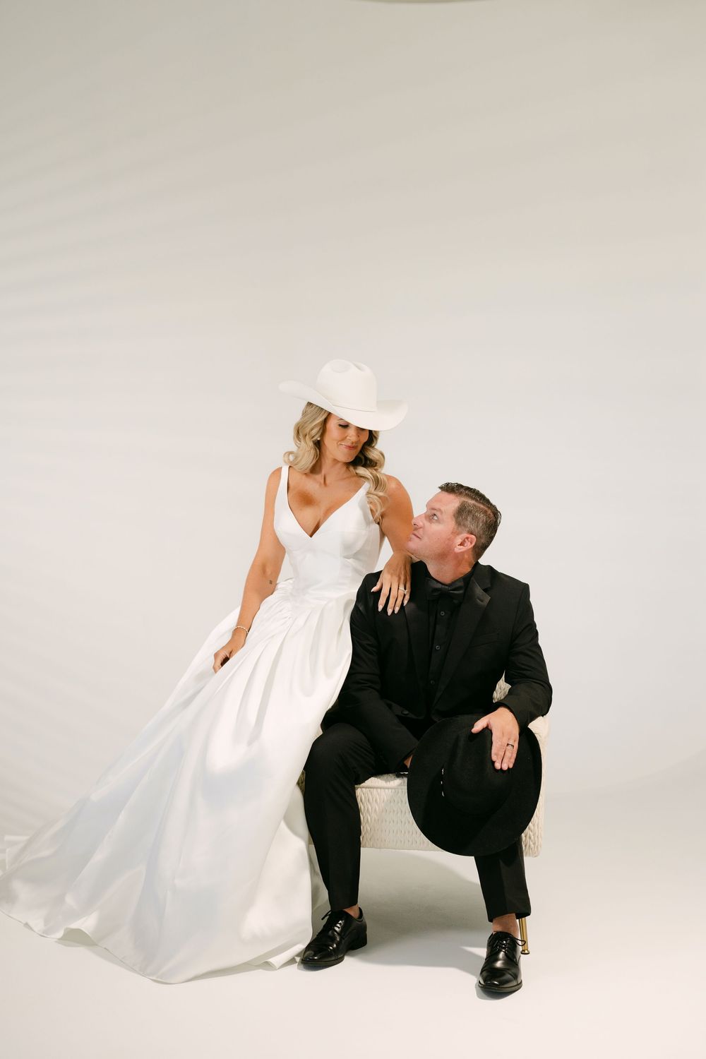 Bride in white gown and cowboy hat poses with groom in black suit holding hat, studio wedding portrait.