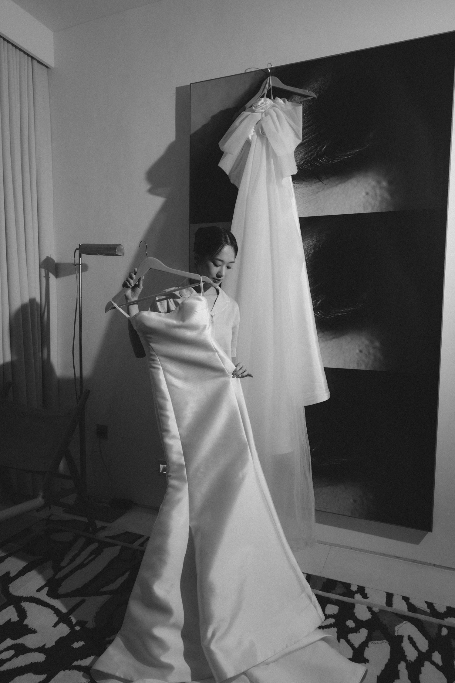 Bride in elegant white satin wedding gown stands beside hanging veil in black and white photo.