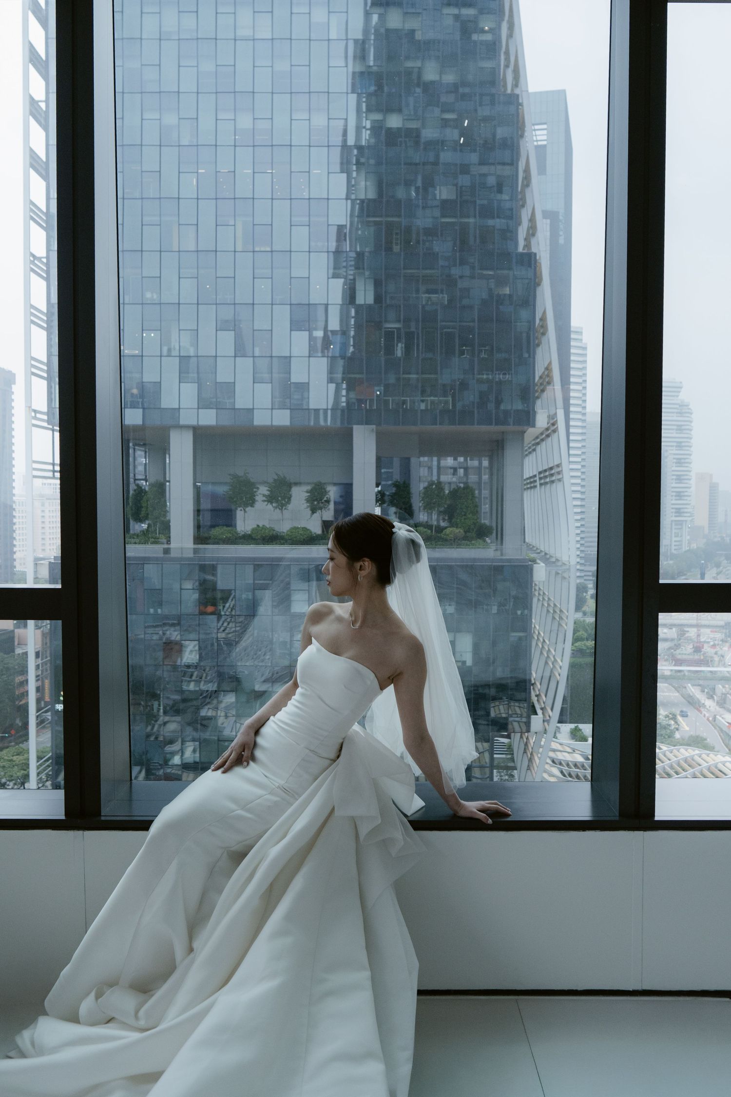 A bride in a flowing white strapless gown and veil sits by a large window overlooking a modern city skyline.