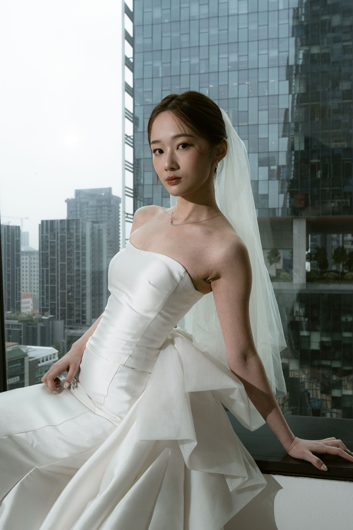 Elegant bride in strapless white gown and veil poses by window with city skyline backdrop.
