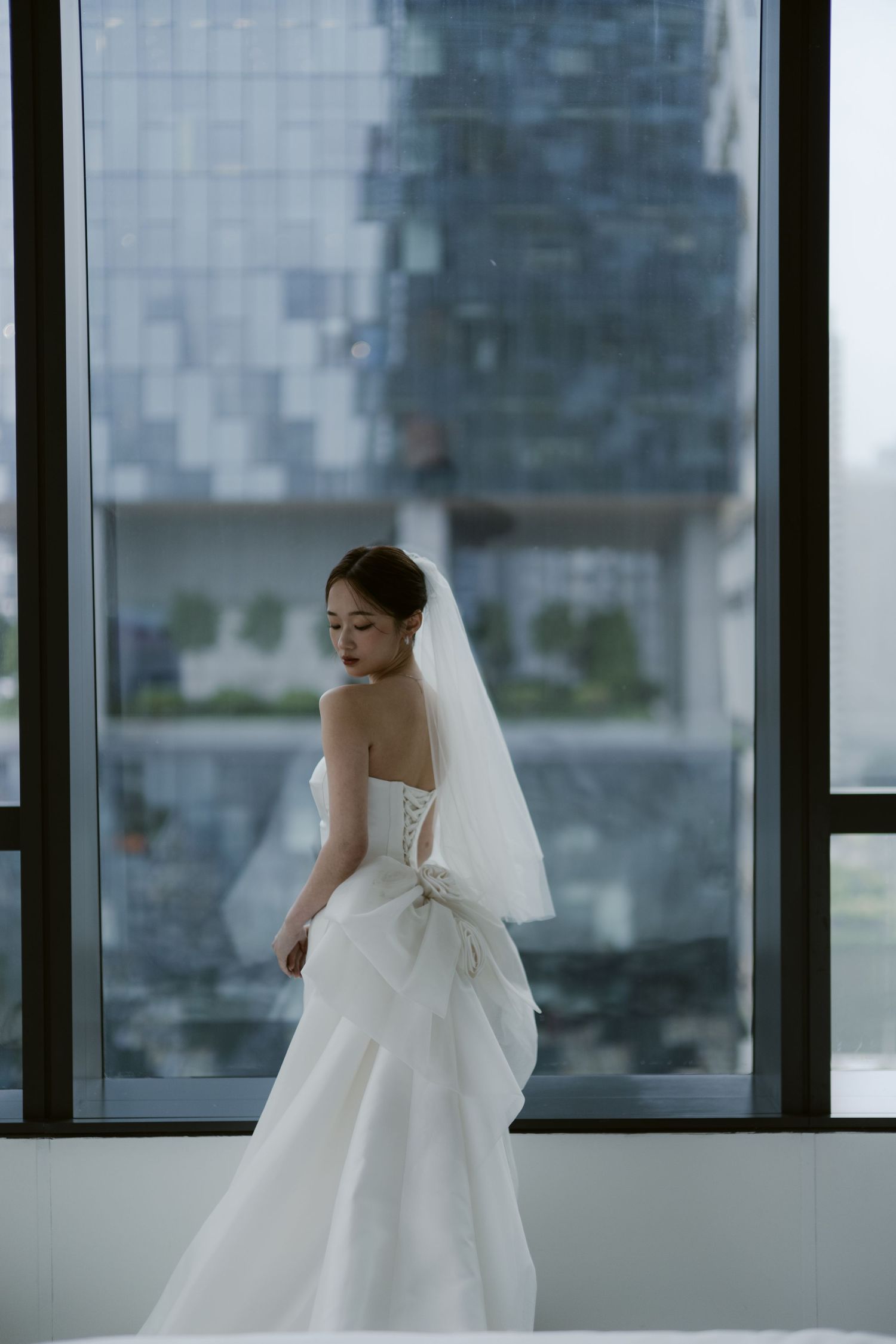 Elegant bride in a strapless white wedding gown with veil and bow, standing by large windows with city view.