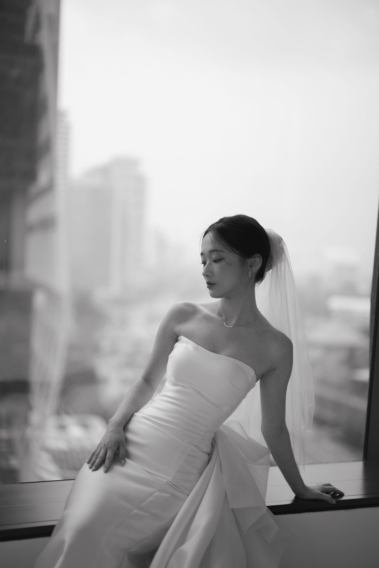 Elegant bride in strapless white gown and veil poses by a large window with city skyline in black and white.