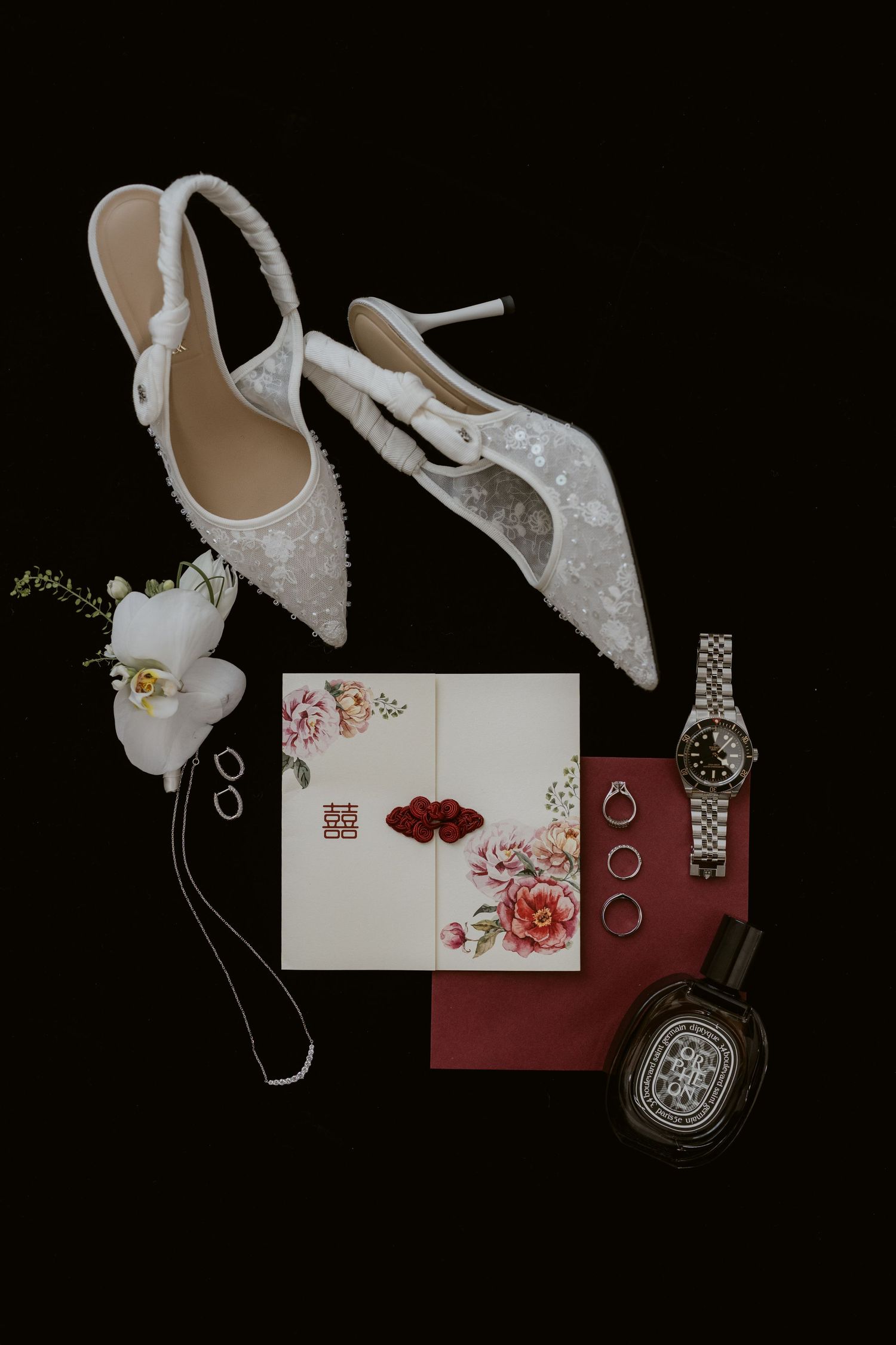Elegant bridal flat lay with white lace heels, floral wedding invitation, silver watch, rings, and perfume on black background.
