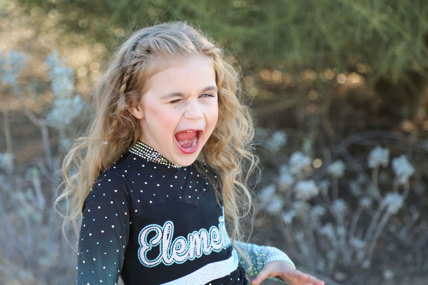 Young girl with long curly hair in a black cheer uniform winks and sticks out her tongue in a playful outdoor portrait.