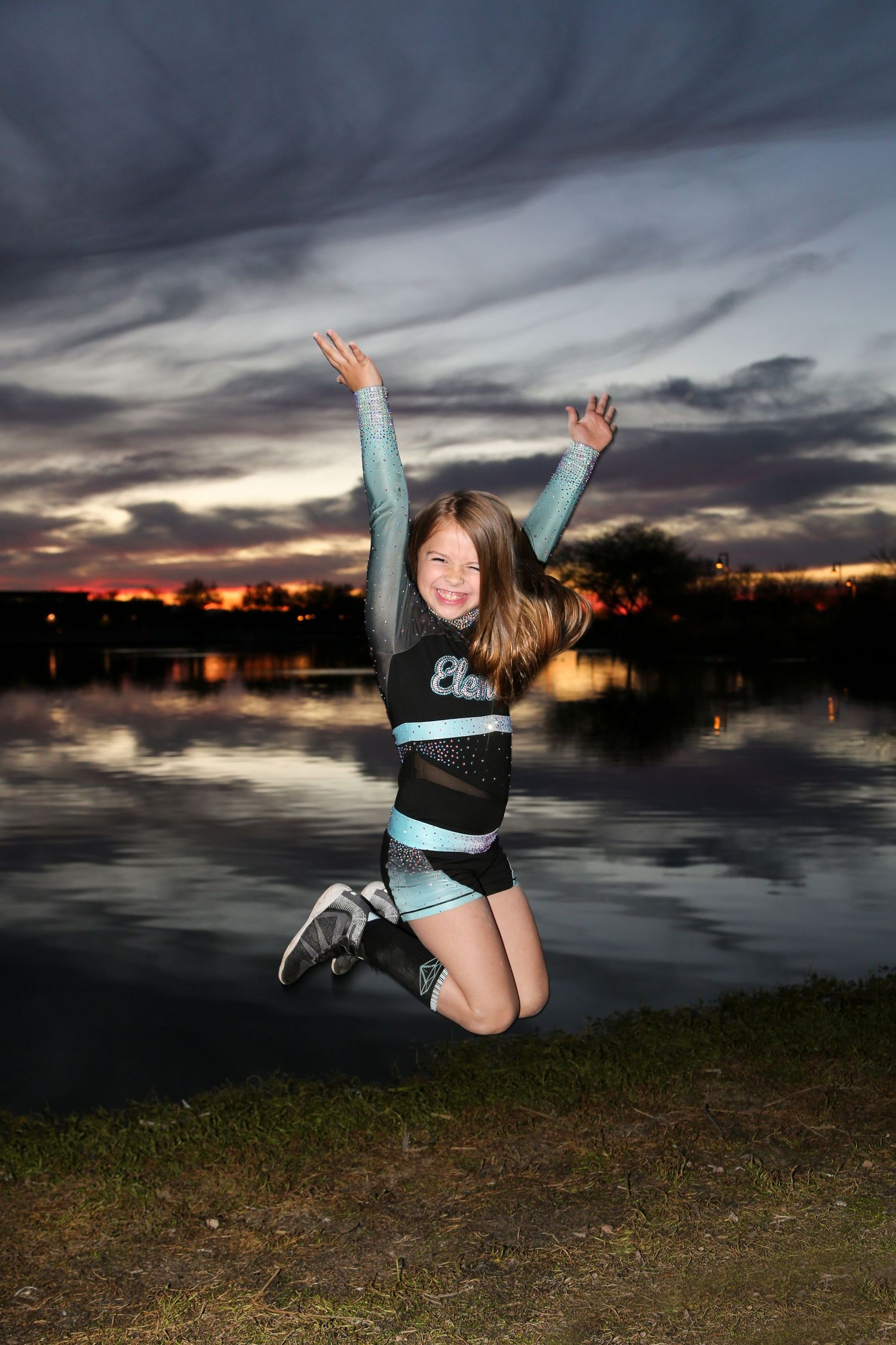 Smiling cheerleader in a teal and black uniform leaps with arms raised beside a reflective lake under a dramatic sunset sky.
