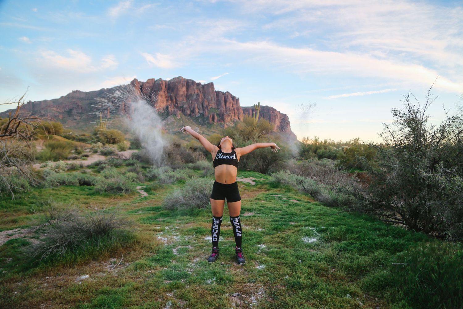 Athlete in black sports gear and knee guards stretches her arms wide on a desert trail with rocky mountains behind her.