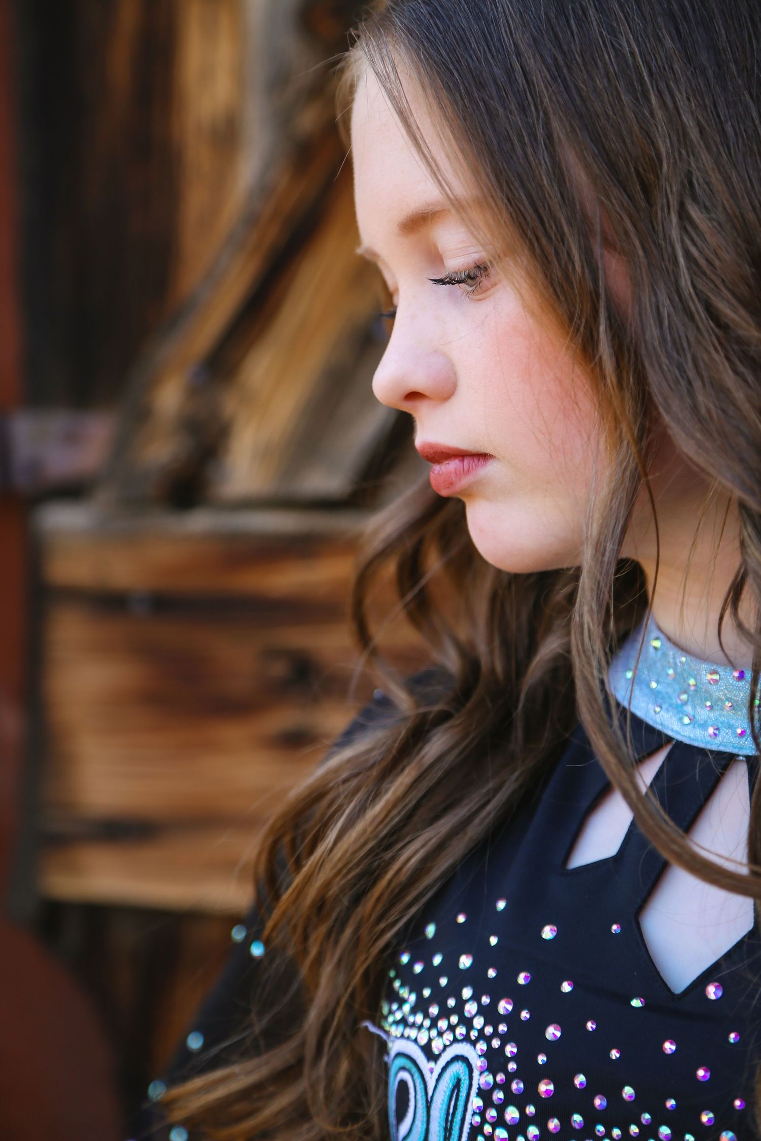 Side profile of a young cheer athlete with long brown hair wearing a black rhinestone top near an aged wooden wall.