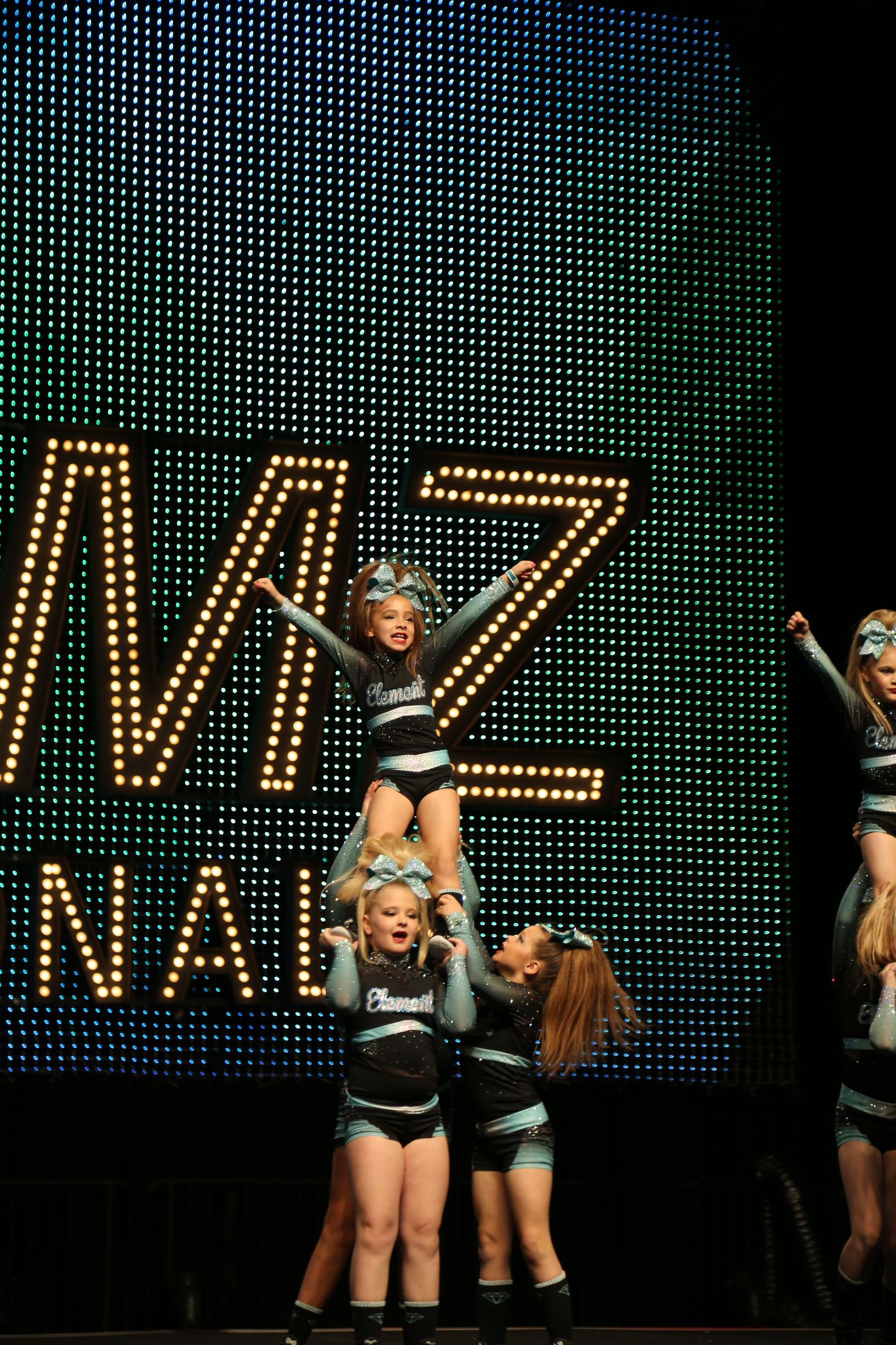 Cheerleaders perform a stunt on stage as a flyer extends her arms above a lit LED display backdrop.