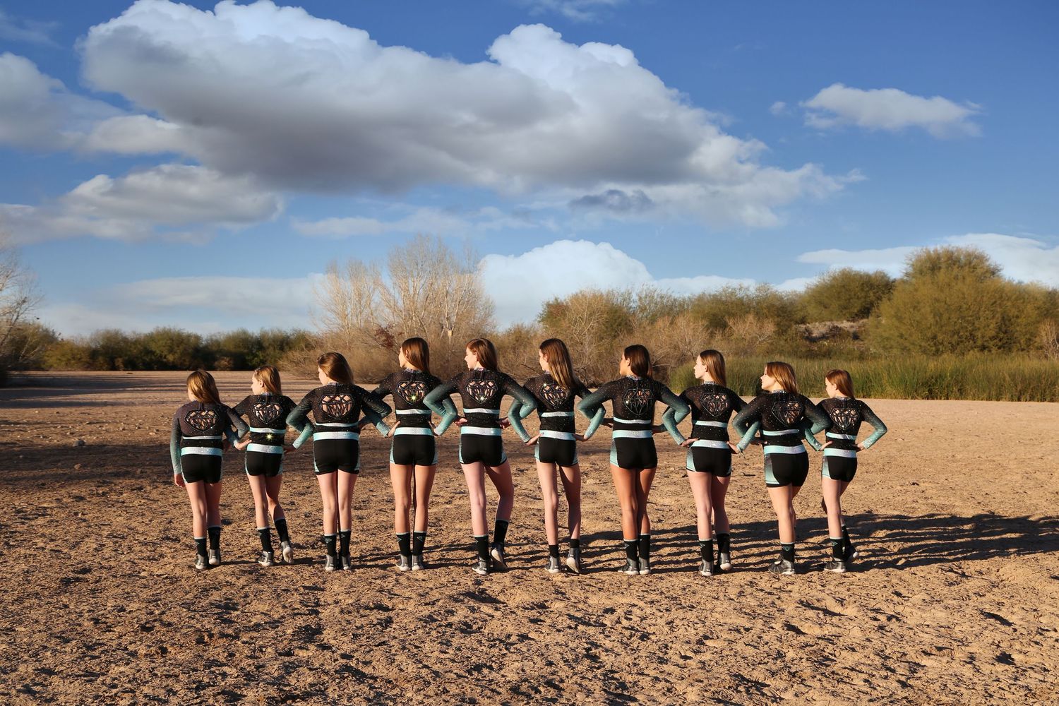 Cheer team stands in a row with arms around each other, facing a desert horizon under a blue sky.
