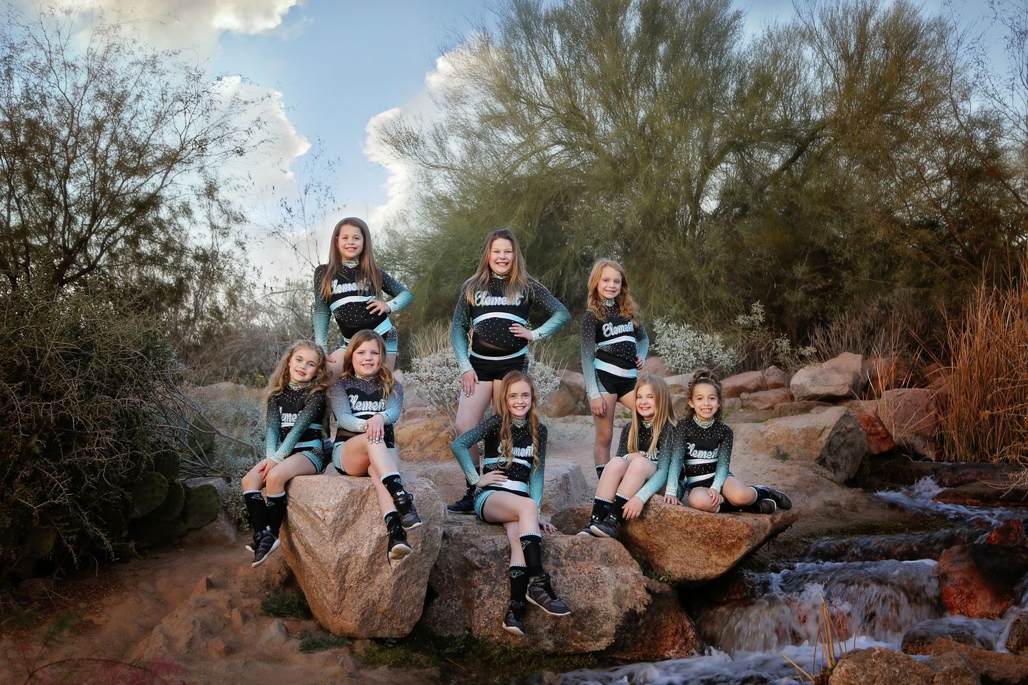 Cheer team in matching teal and black uniforms poses on rocks beside a small desert stream at sunset.